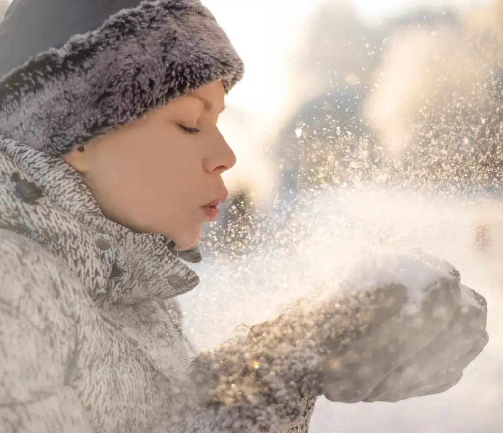 a woman blowing snow in her hands implying she has experienced the benefits of winter wellness tips for a healthy season